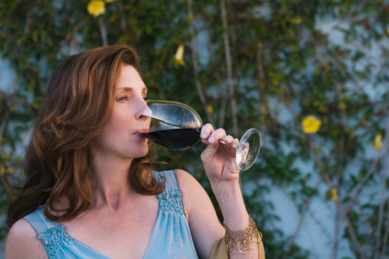 Uma mulher de cabelos castanhos e vestido azul, bebendo vinho em uma taça.