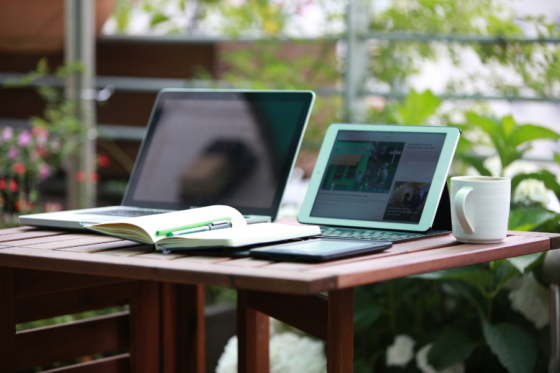 Mesa de madeira com notebook e tablet em ambiente externo.